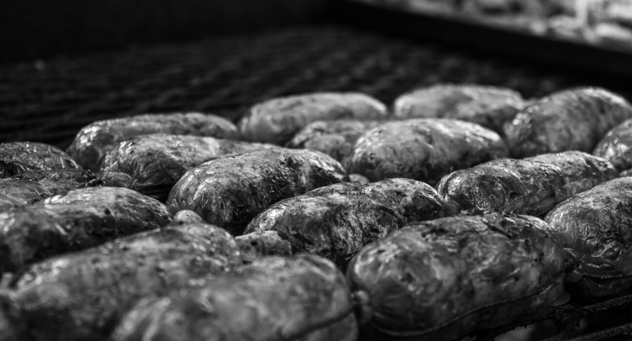 Black and white close-up image of sausages being grilled over an outdoor barbecue pit.