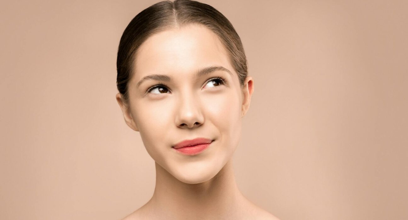 A young woman with radiant skin looks upward against a neutral background, embodying natural beauty.