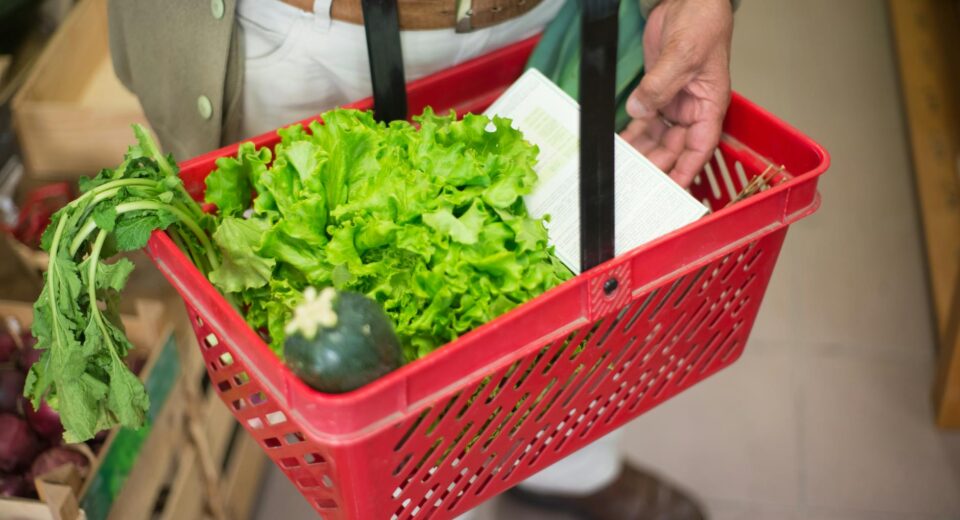 Close-up of a hand carrying a shopping basket filled with fresh greens in a market.
