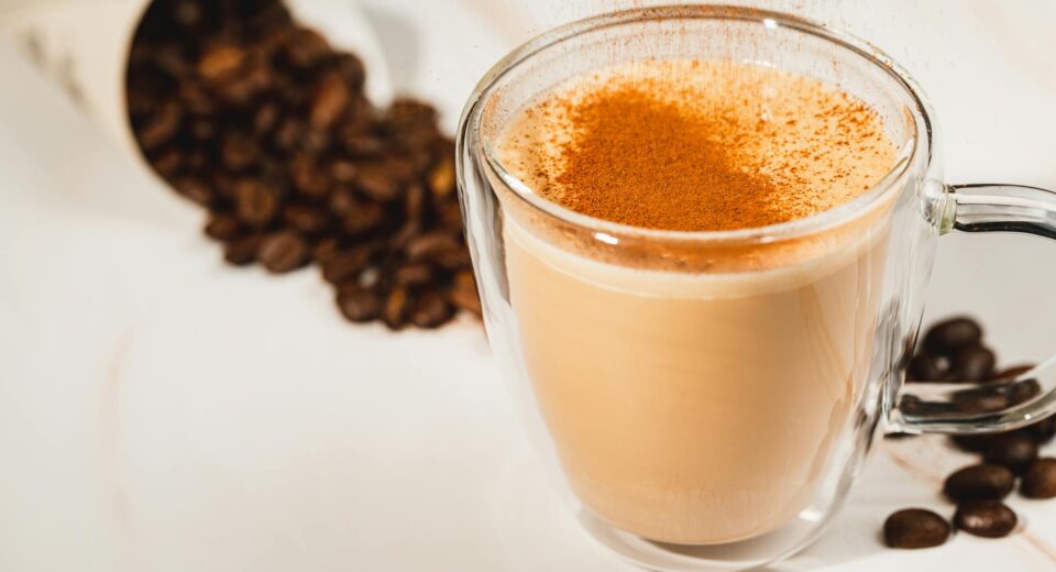 A warm latte in a glass cup topped with cinnamon, surrounded by coffee beans.