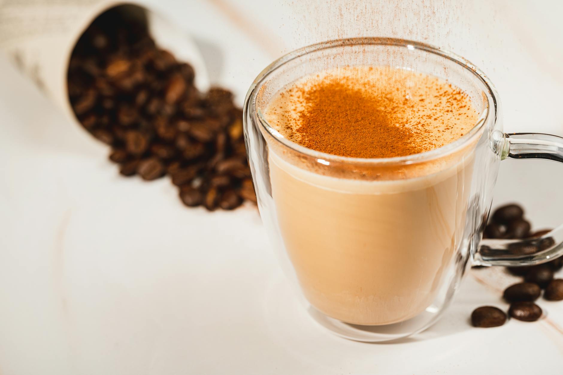 A warm latte in a glass cup topped with cinnamon, surrounded by coffee beans.