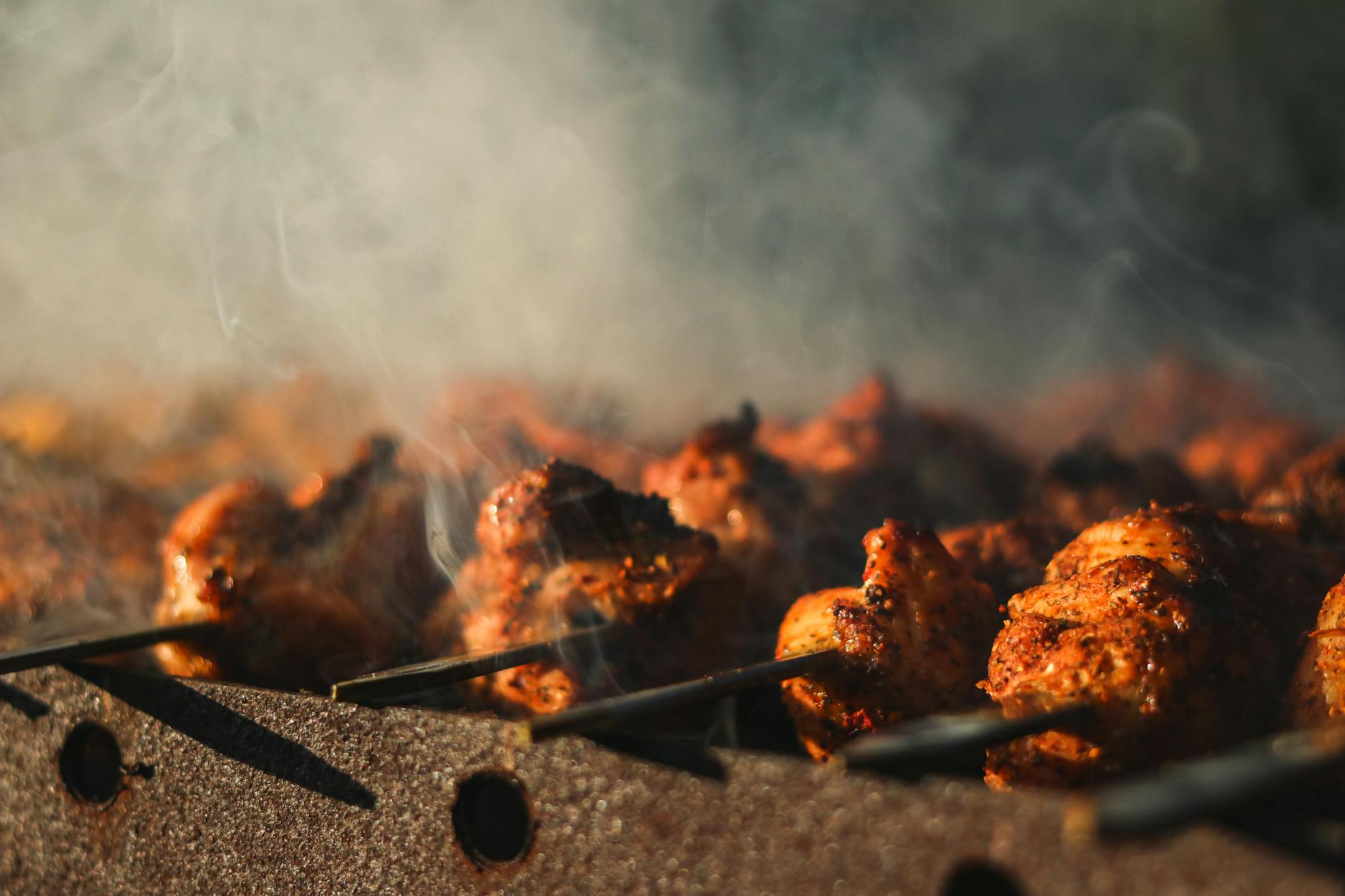 Close-up of skewered grilled chicken sizzling with smoke on barbecue.
