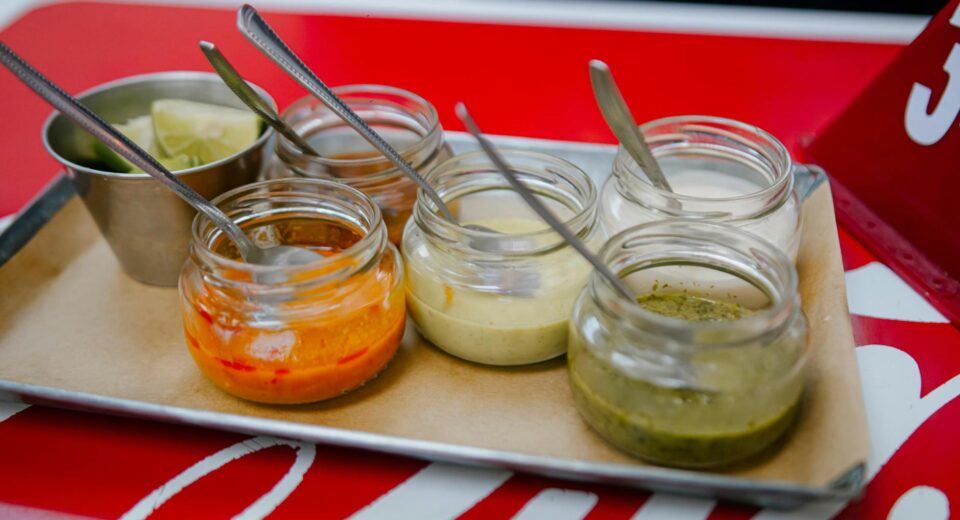 Close-up of colorful sauces in glass jars with spoons on a tray, perfect for culinary presentations.