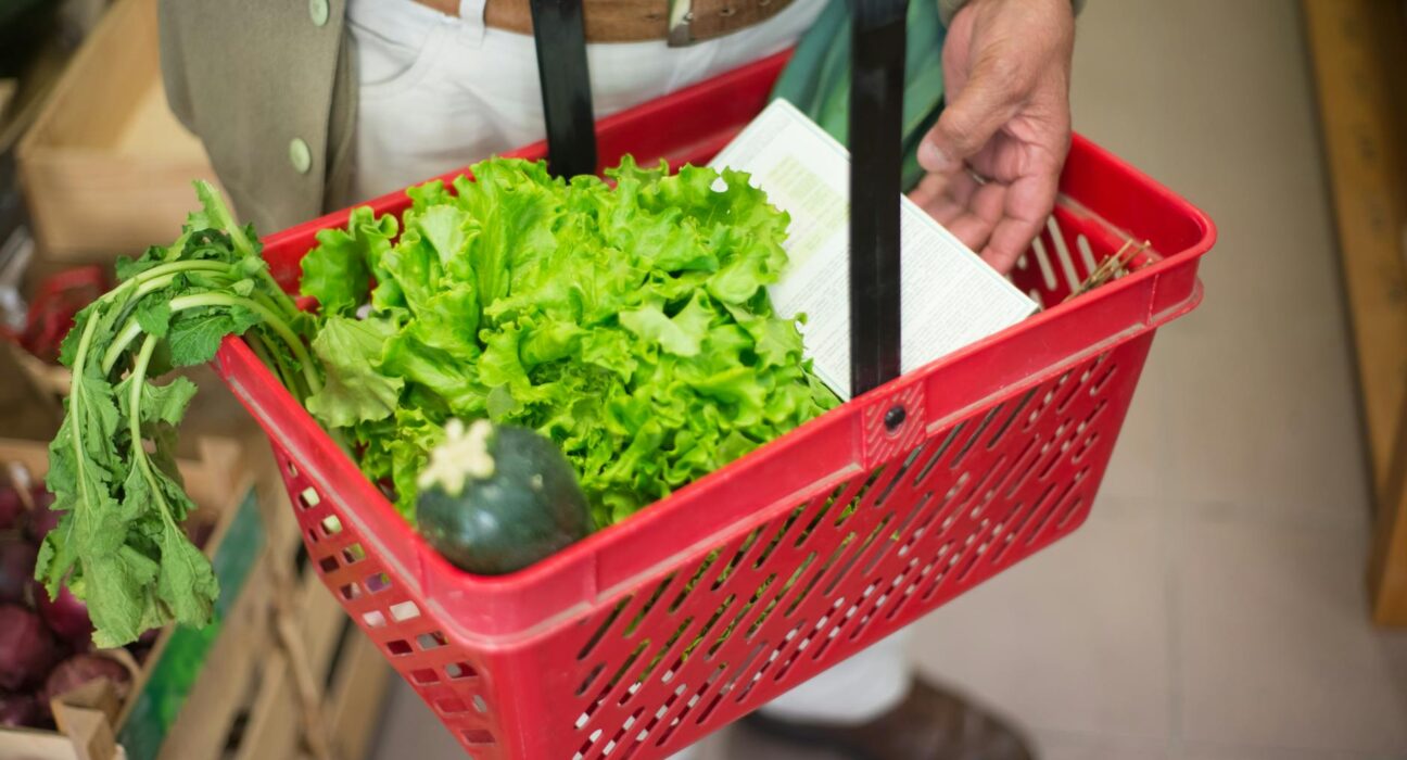 Close-up of a hand carrying a shopping basket filled with fresh greens in a market.