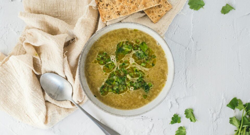 A bowl of fresh herb soup served with crackers and a spoon on a light background.
