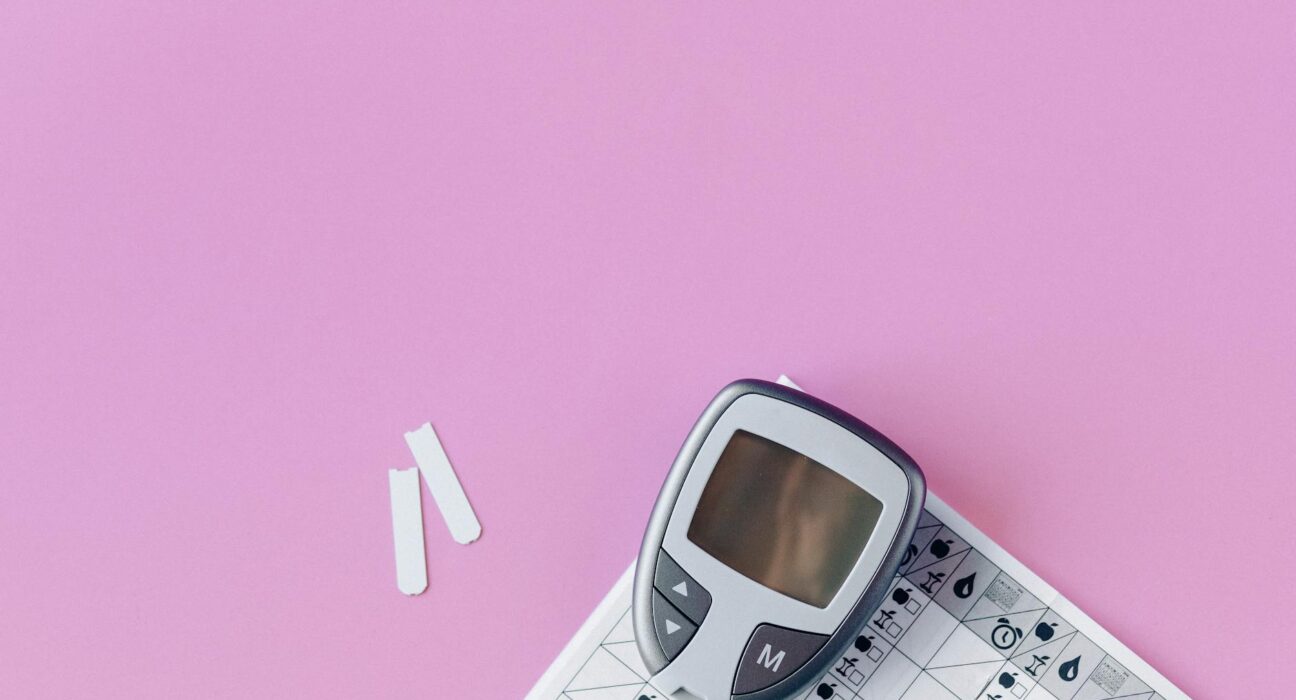 Flat lay of a blood glucose monitor and test strips on a pink surface.