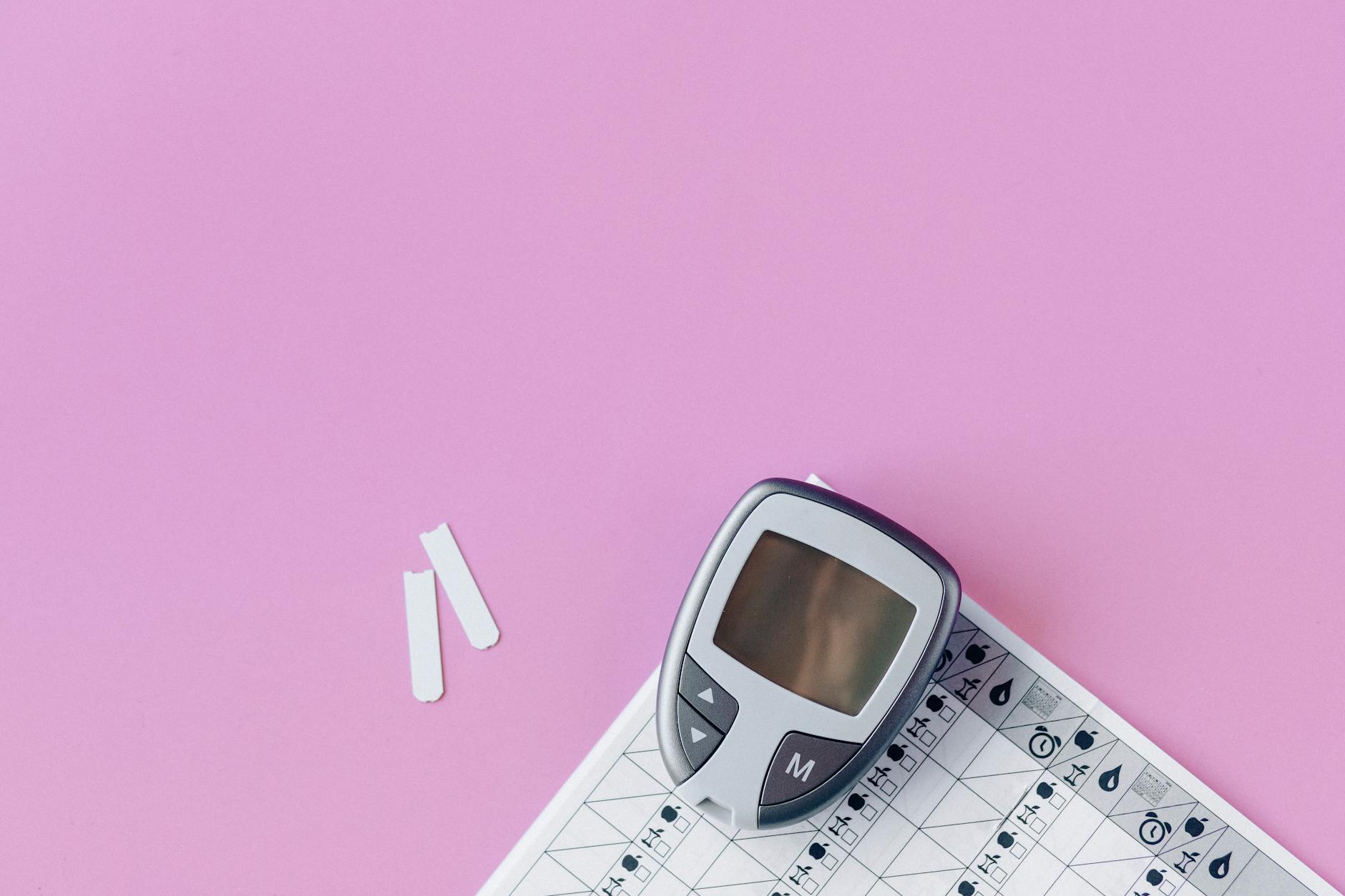 Flat lay of a blood glucose monitor and test strips on a pink surface.