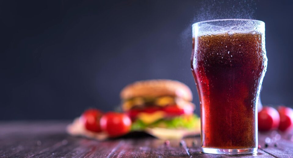 A close-up of a fizzy drink with a hamburger and tomatoes in the background, perfect for fast food themes.