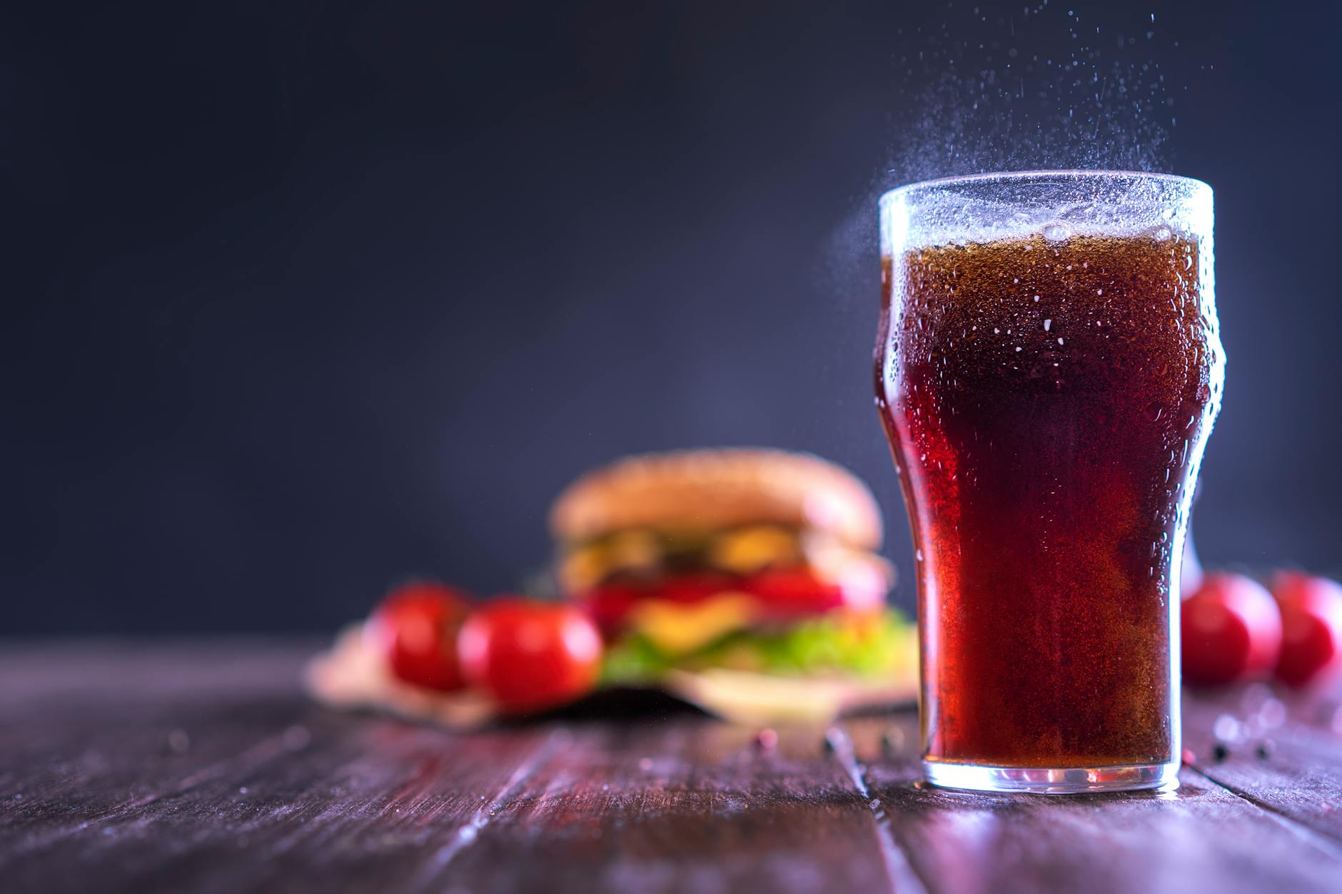A close-up of a fizzy drink with a hamburger and tomatoes in the background, perfect for fast food themes.