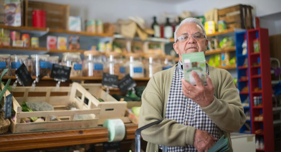 Elderly man examines product in grocery store aisle, representing daily shopping routine.
