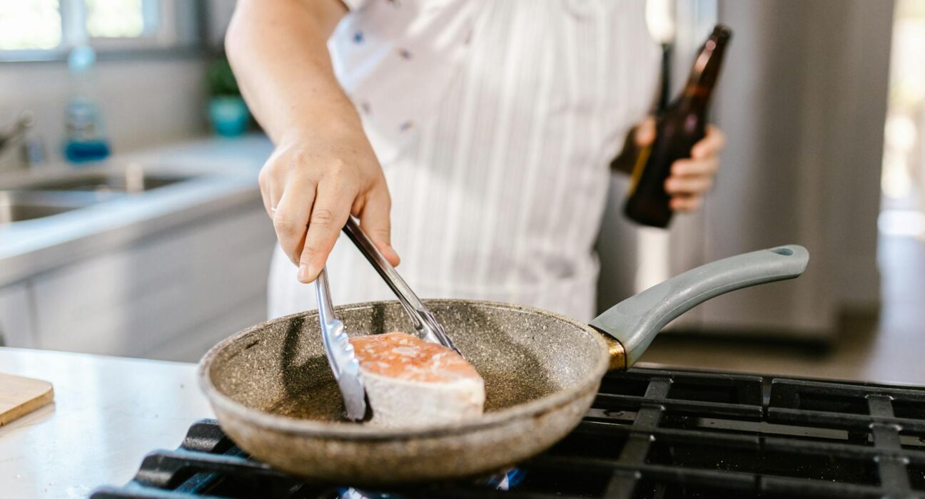 A person cooking salmon fillet in a frying pan on a kitchen stove.