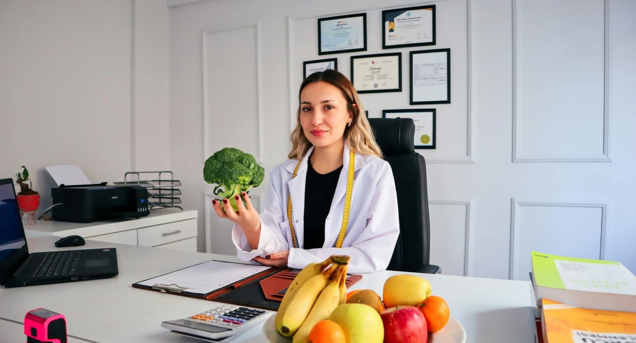 Female nutritionist in office holding broccoli, surrounded by fruits, promoting healthy lifestyle.