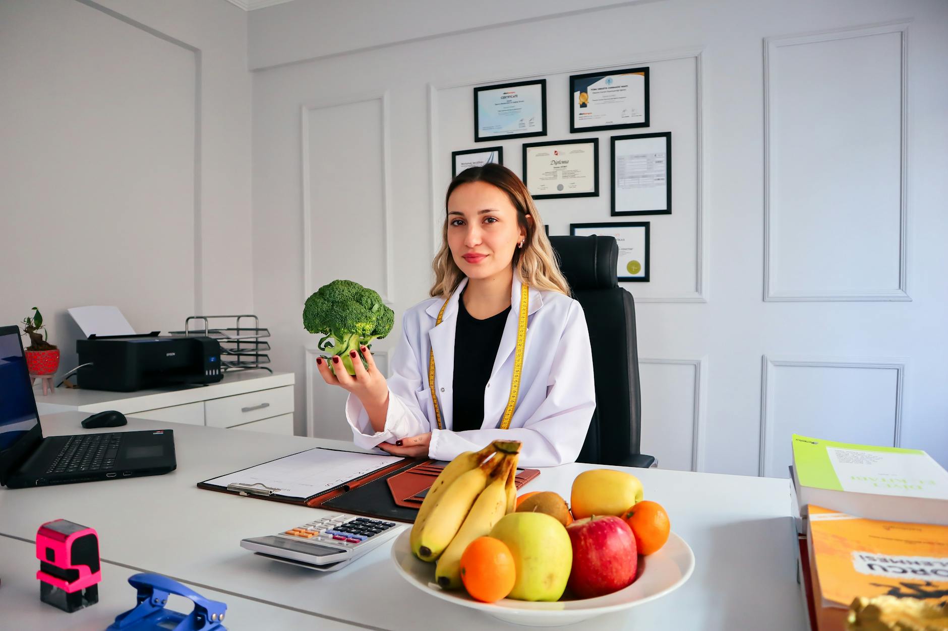 Female nutritionist in office holding broccoli, surrounded by fruits, promoting healthy lifestyle.