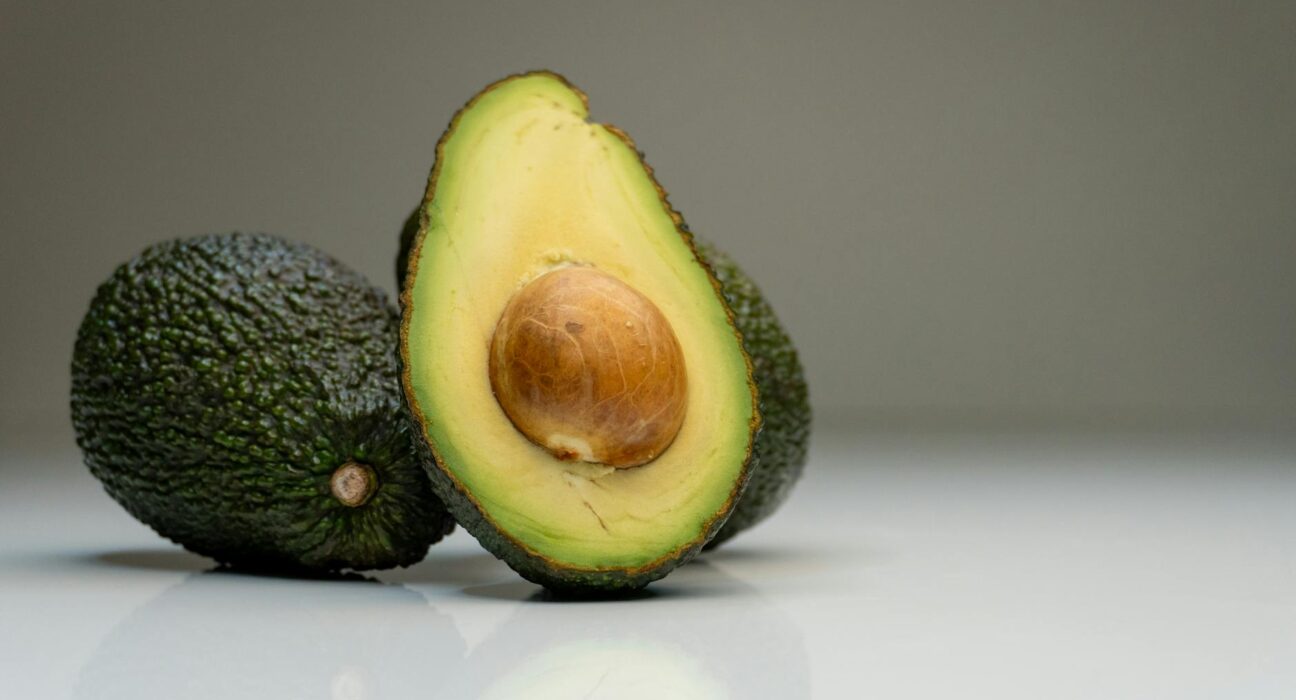 Close-up of ripe avocados with a sliced half showing the seed, against a gray background.