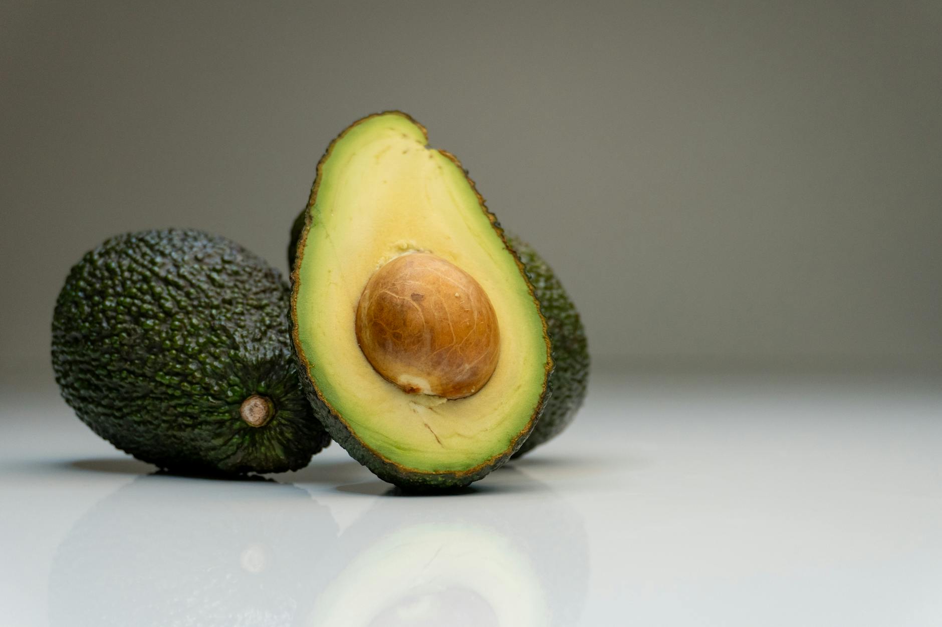 Close-up of ripe avocados with a sliced half showing the seed, against a gray background.
