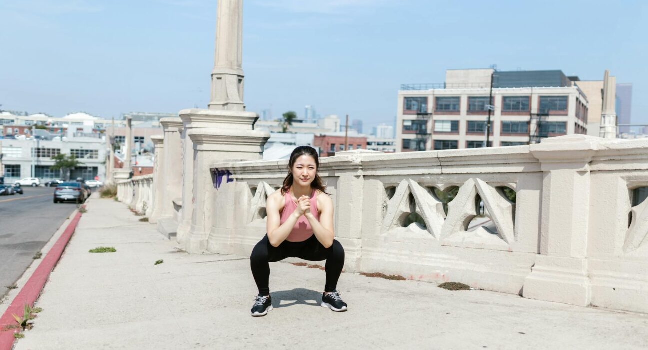 Woman exercising outdoors, performing squats on a city sidewalk.