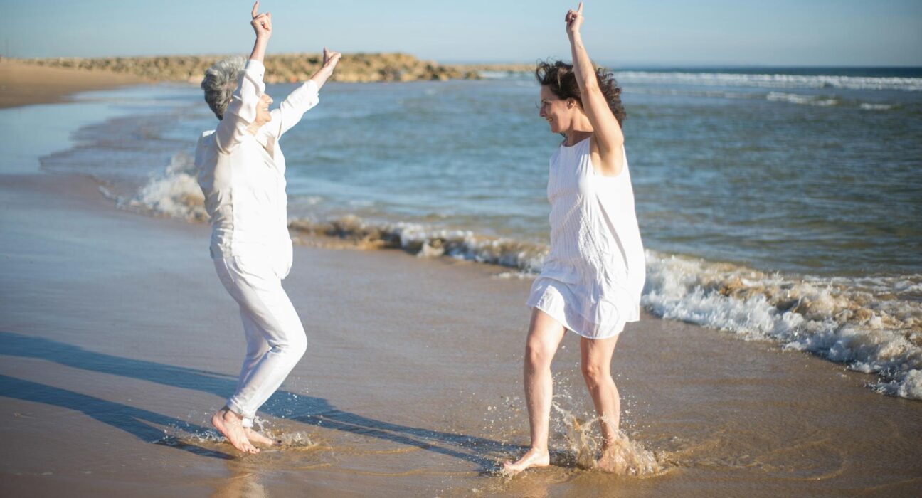 Two women laughing and dancing on a sunny beach, enjoying a carefree day by the ocean.
