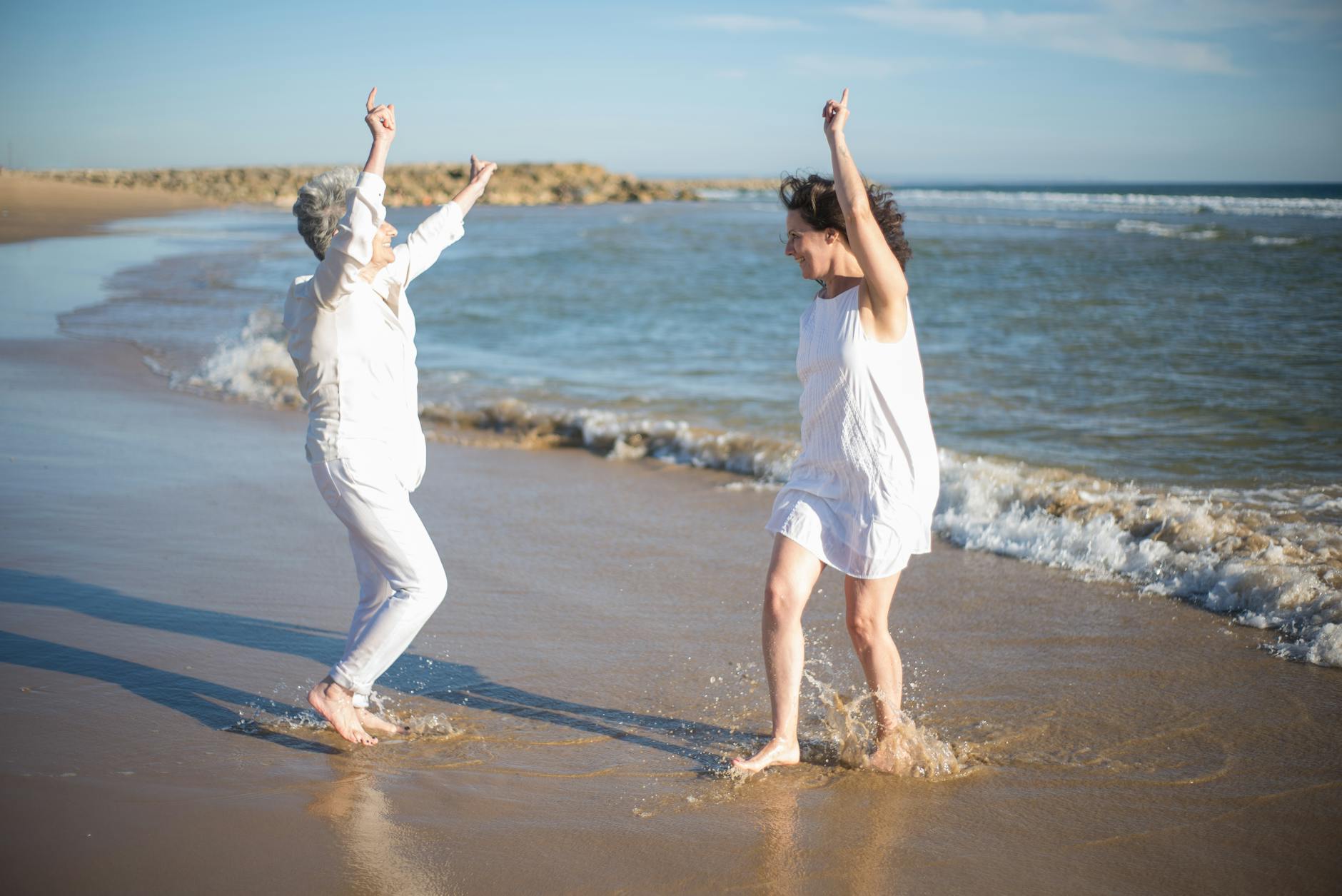 Two women laughing and dancing on a sunny beach, enjoying a carefree day by the ocean.