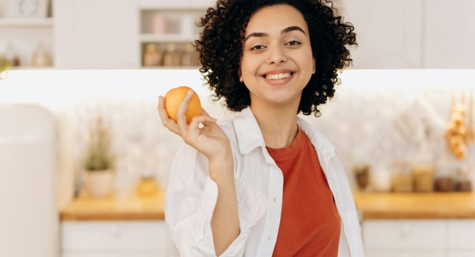 A joyful woman with curly hair holding an orange in her kitchen, promoting a healthy lifestyle.
